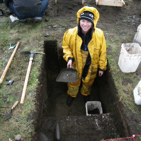 Shannon Tushingham stands in a dug-out section of earth while wearing a yellow raincoat, holding a digging tool during an archaeological dig. 