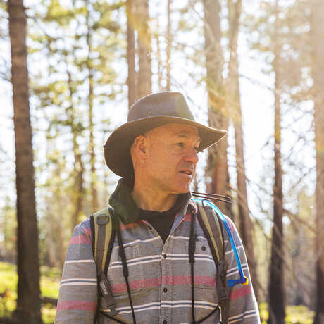Jack Dumbacher stands backlit against a thick swath of trees in Caples Creek, wearing a striped button up shirt and a wide brimmed hat with binoculars. 