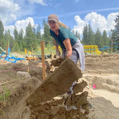 Shannon Tushingham digs up a clump of dirt during an archaeological dig and tosses it at the camera,with the dirt coming close to the viewer. 