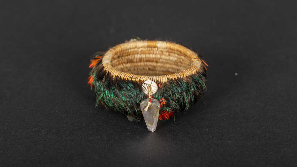 A green, white, and red feathered pomo basket with shells on the outside sits against a black backdrop. 